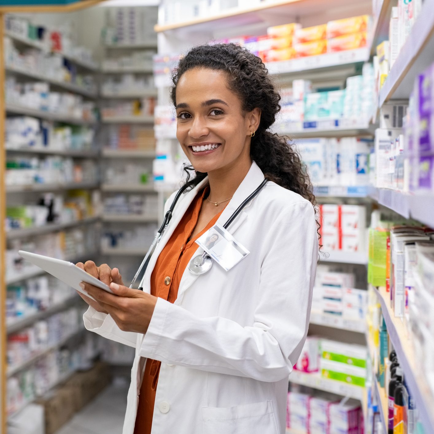 Happy friendly multiethnic pharmacist doing inventory in a provided and modern pharmacy while looking at camera. Portrait of smiling young doctor woman working in drugstore with digital tablet. African smiling druggist working at hospital pharmacy.